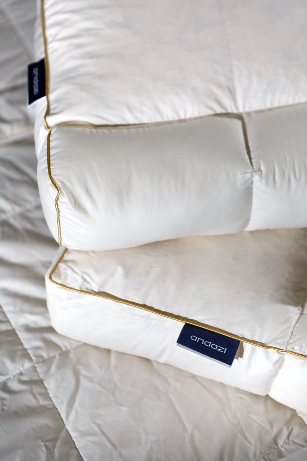 Close-up of two thick white pillows with gold piping on a bed, featuring Andazi tags