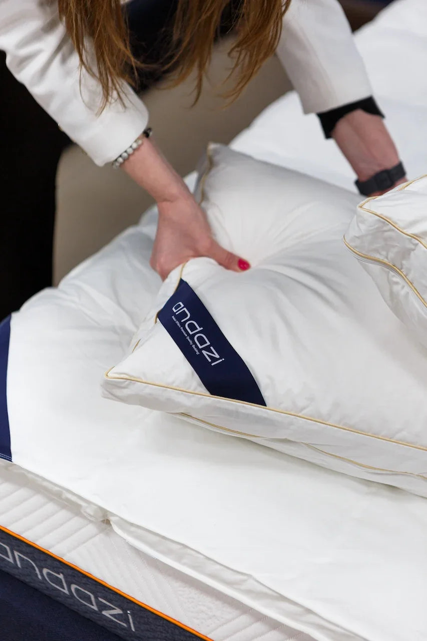 Person arranging a white Andazi pillow on a bed with mattress, close-up view