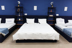 Showroom display of three modern beds with white bedding and pillows against a blue wall