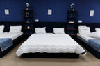 Showroom display of three white mattresses with pillows on dark bed frames against a blue wall