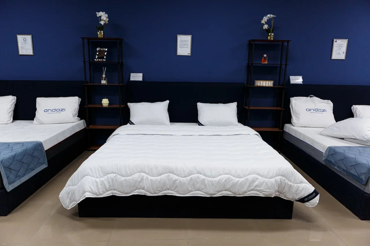 Showroom display of three white mattresses with pillows on dark bed frames against a blue wall